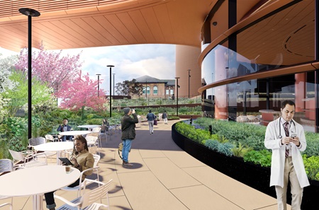 A design rendering shows people enjoying an outdoor patio seating area with round tables and chairs in front of the Pavilion and underneath the connector bridge crossing 34th Street. A thick area of landscaping hides the street from view.